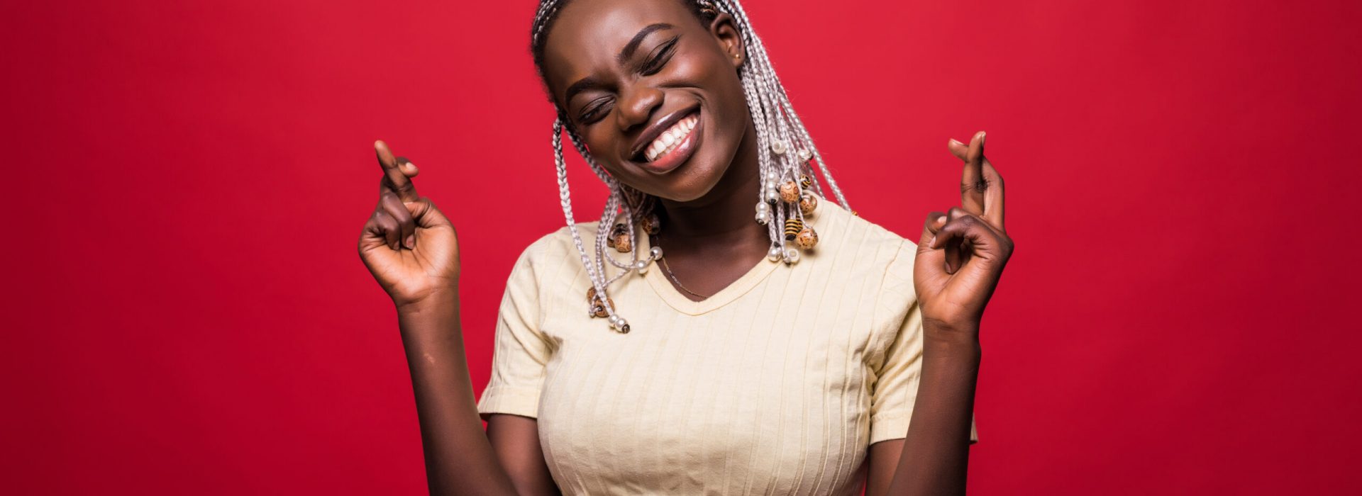Beautiful young Afro American woman is holding fingers crossed and smiling, isolated on red background